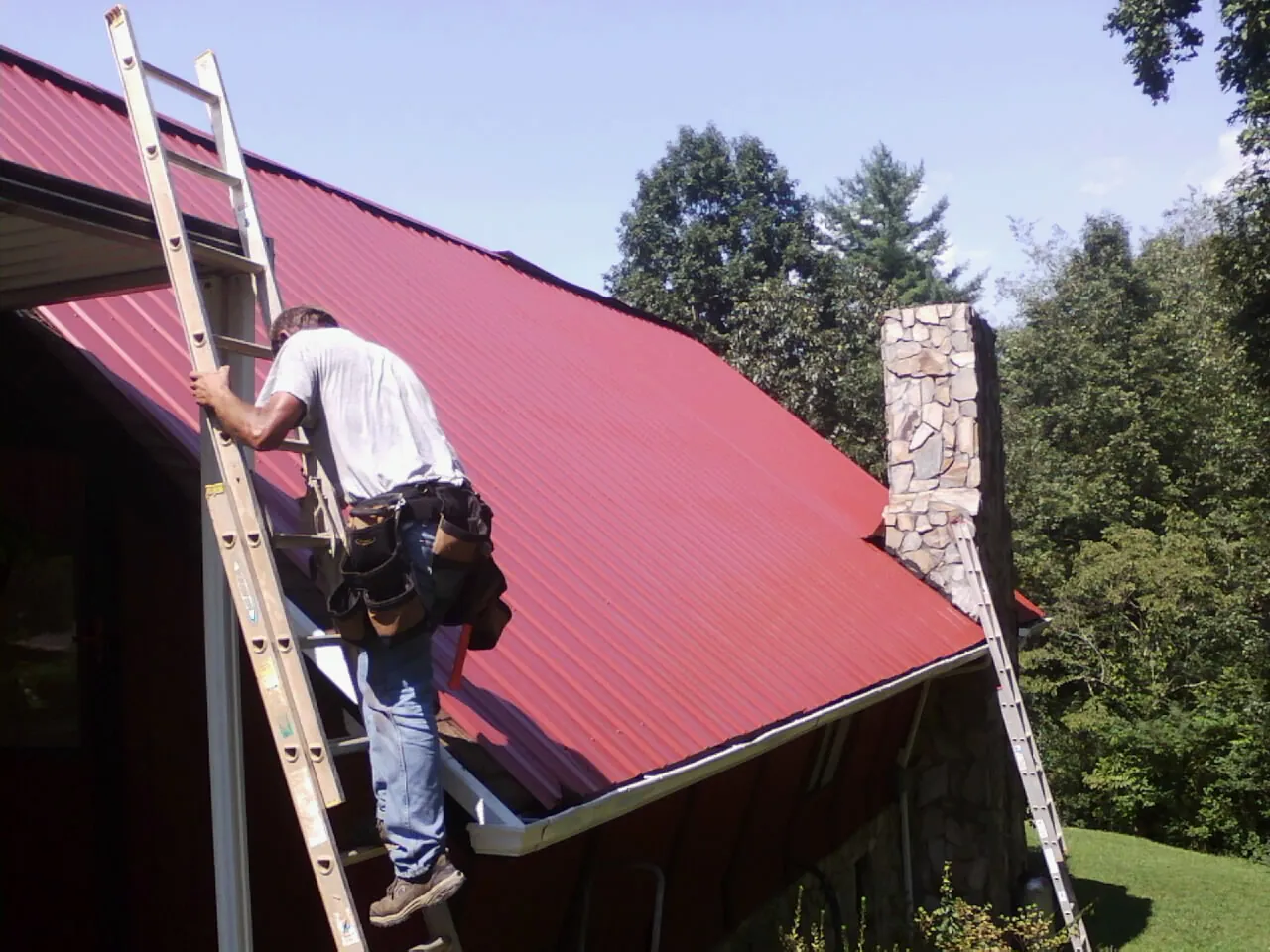 Roofer on a ladder beside a red metal roof and stone chimney — Soffit & Fascia Repair by Roofing Brothers in Warrenville